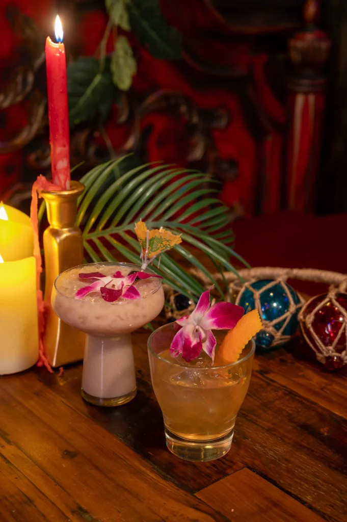 Two orchid-garnished cocktails on a wooden table beside candles and greenery.