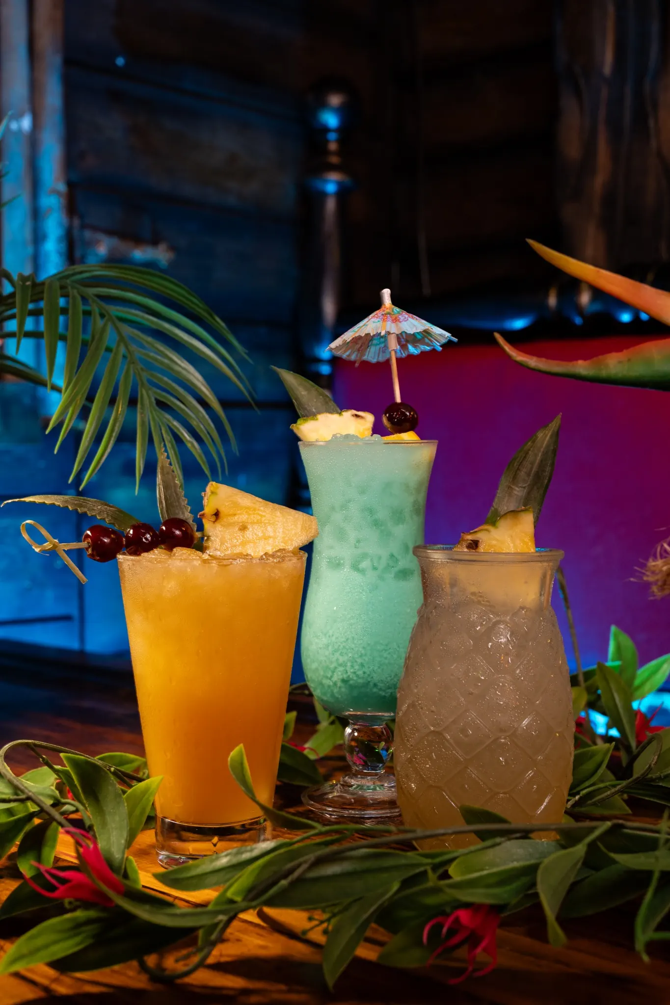 Three tropical cocktails with pineapple garnishes surrounded by lush green foliage.